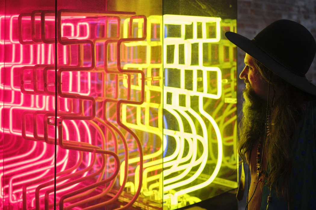 A neon work in four different colors casts a green-yellow light on a person's face who stands close to the work.